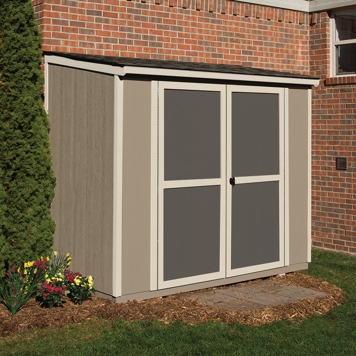 A beige shed with double doors and a lock, located near a brick wall with flowers and mulch in the foreground.  A beige shed with double doors and a lock, located near a brick wall with flowers and mulch in the foreground.