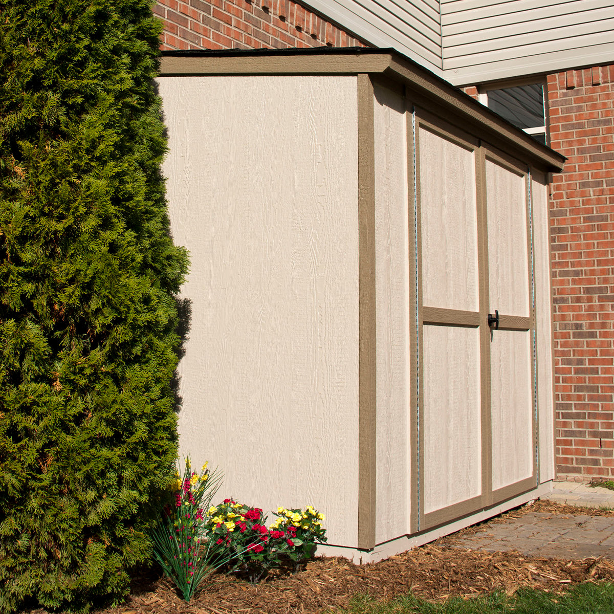 A white shed with tan trim and double doors, situated beside a tree and flowers, against a brick wall in the background. A white shed with tan trim and double doors, situated beside a tree and flowers, against a brick wall in the background.