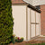 A white shed with tan trim and double doors, situated beside a tree and flowers, against a brick wall in the background. A white shed with tan trim and double doors, situated beside a tree and flowers, against a brick wall in the background.