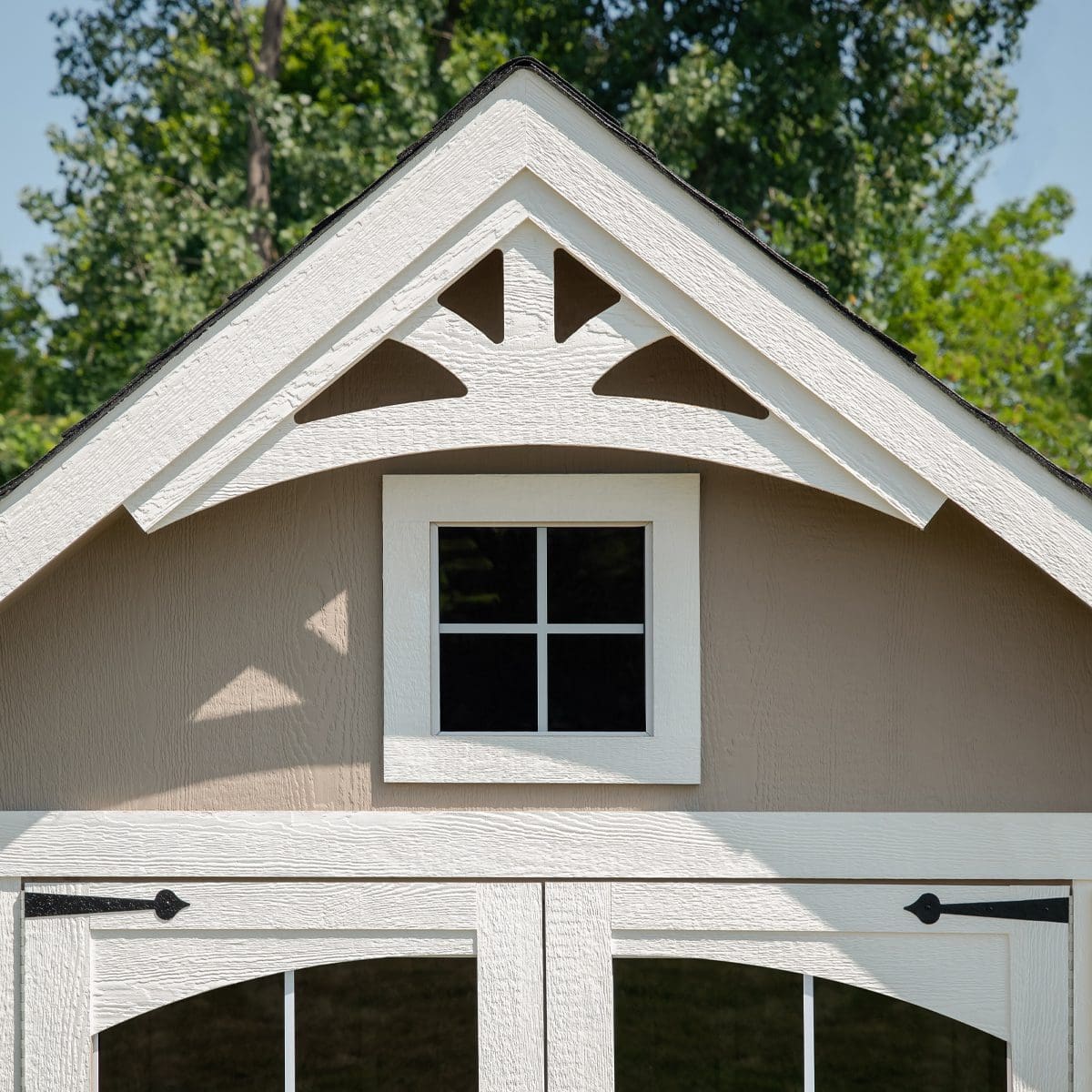 Close-up of a shed with decorative white trim, a small window, and a curved roof gable. The background features greenery.