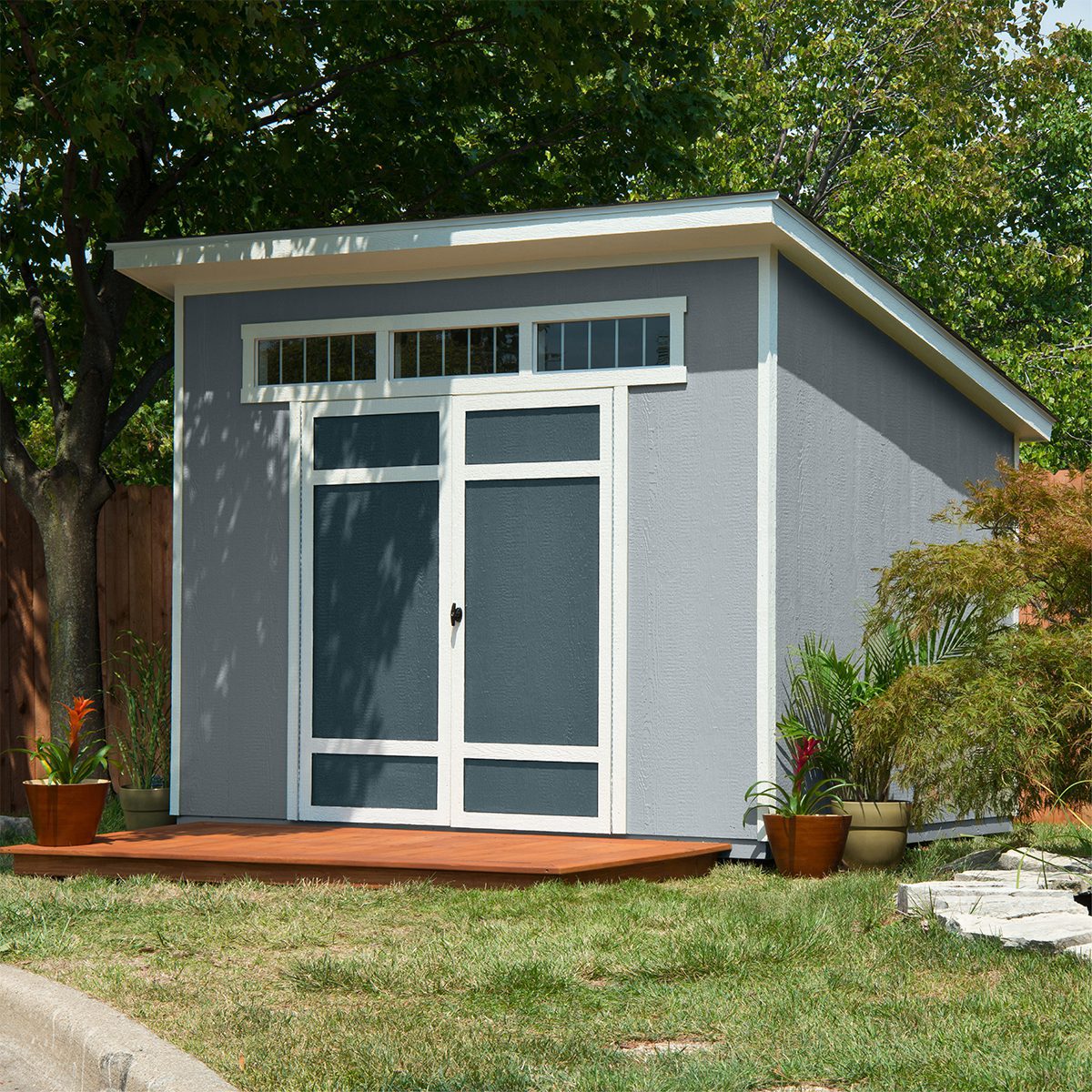 Modern backyard shed with transom windows, double doors, and a slanted roof, surrounded by plants and a wooden deck.