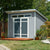 Modern backyard shed with transom windows, double doors, and a slanted roof, surrounded by plants and a wooden deck.