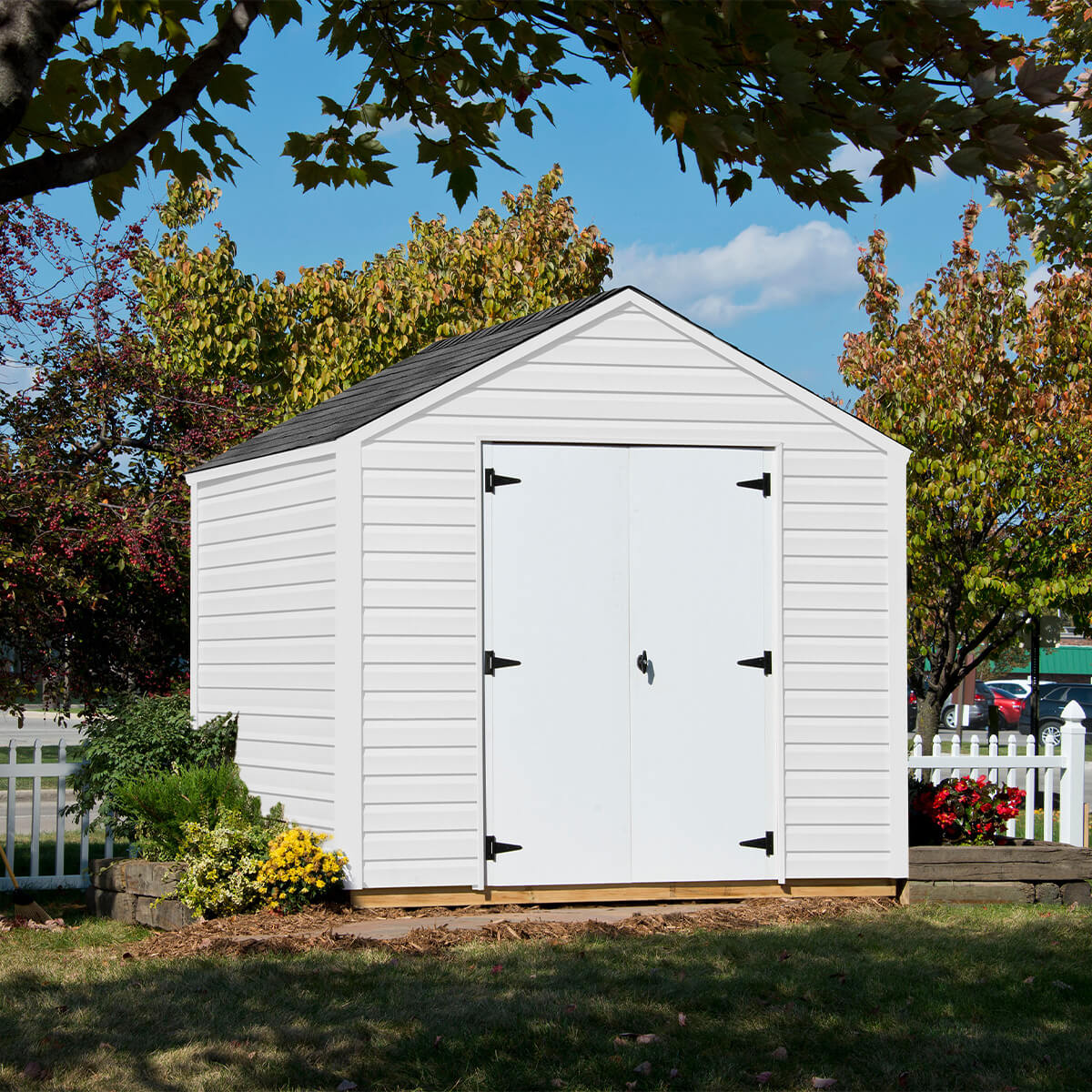 A shed with double doors, black hardware, surrounded by trees and flowers, with a white picket fence in the background.