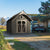A dark gray shed with a pitched roof, decorative window, and double doors, located on a dirt path with a truck and mountains in the background.